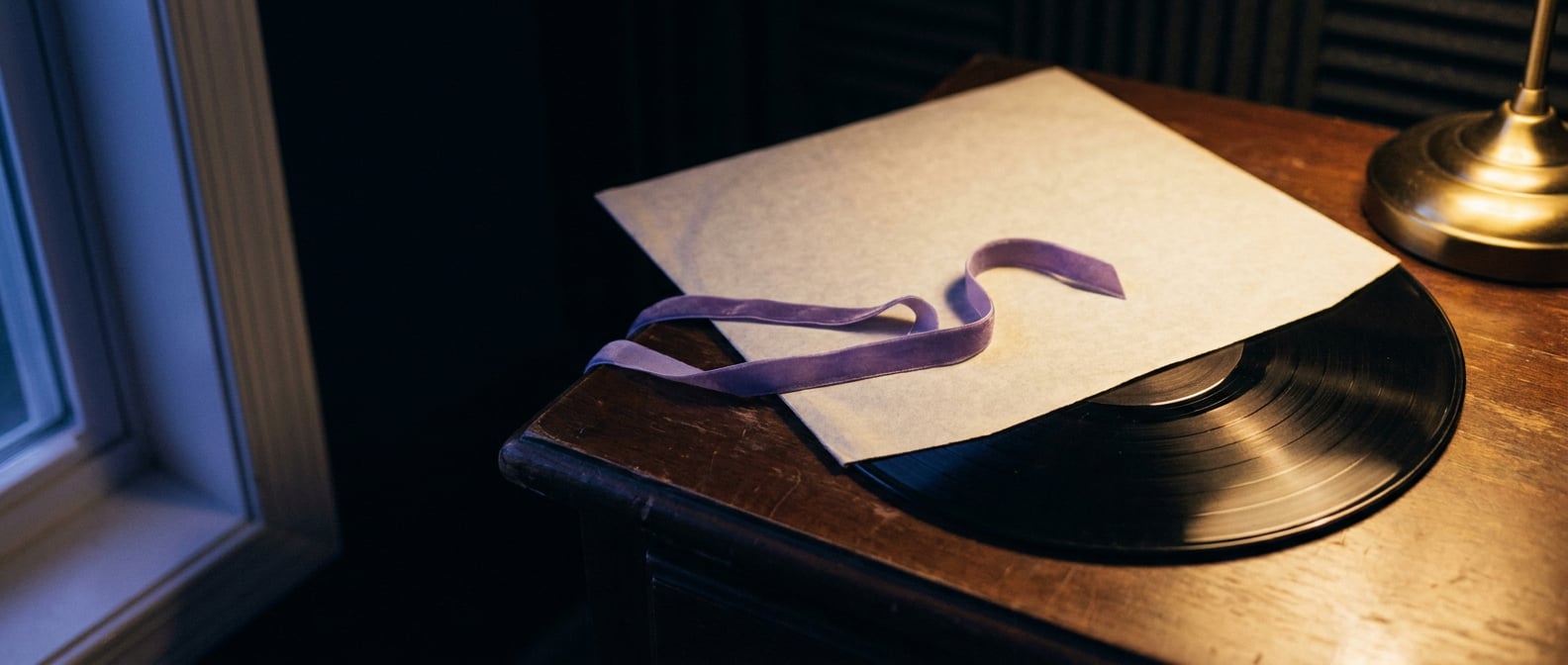 Vinyl halb aus seiner Papierhülle gezogen, ein Lavendel-Bändchen über die Hülle drapiert, gedämpftes Indigo-Licht aus dem Fenster links, warmer Lichtpool aus einer Messinglampe rechts.
