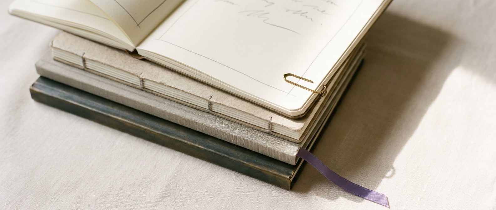 Stapel fadengebundener Notizbücher auf cremefarbenem Leinen, oberstes aufgeschlagen mit handschriftlichen Bleistift-Notizen, ein lavendelfarbenes Seidenband zieht sich aus dem unteren Buch heraus.