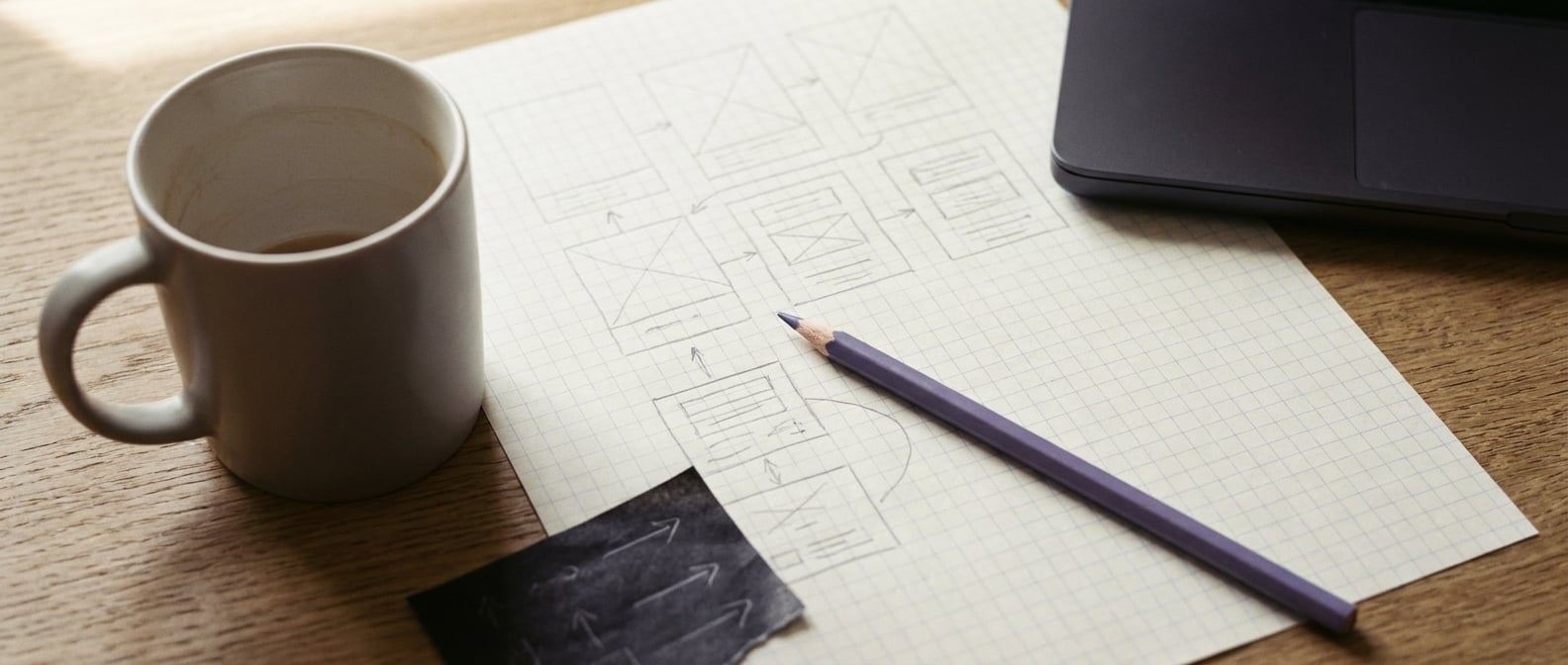 Wireframe-Skizzen mit Bleistift auf Karopapier, daneben Porzellan-Tasse, Lavendel-Bleistift quer abgelegt, Carbonpapier-Ecke, angeschnittener geschlossener Laptop.